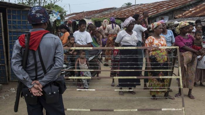 A crowd of people stand behind a fence while an armed officer watches them